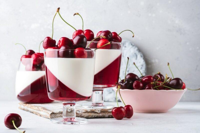 Three glasses on a table, each with a layer of gelatin and one of white panna cotta, with cherries on top, next to a bowl of more cherries