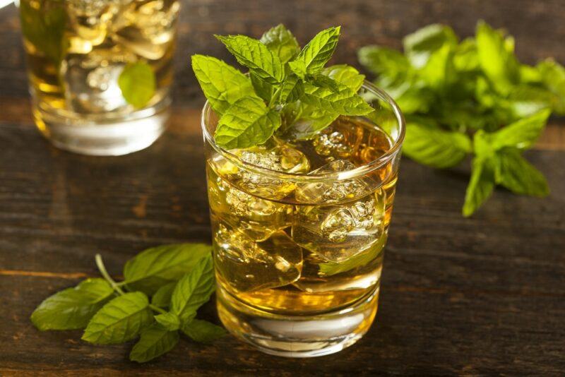 A glass containing a mint julep cocktail with ice, garnished with mint, with more mint on the table