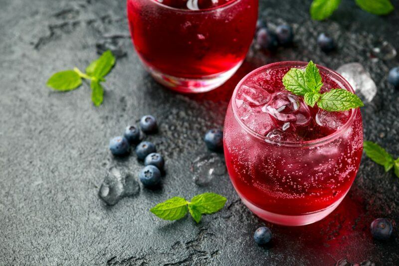 A black table with blueberries and mint leaves, next to two glasses of a French Quarter blueberry smash cocktail