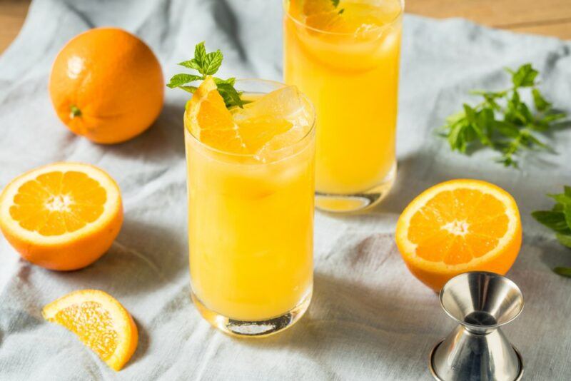 Two glasses containing a Screwball screwdriver cocktail on a white cloth, surrounded by orange segments and herbs