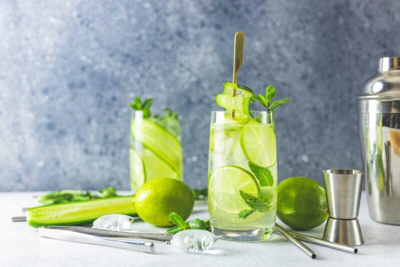 Two tall glasses of a Southern Comfort and Mountain Dew in front of a grey wall, with cucumber, lime, ice