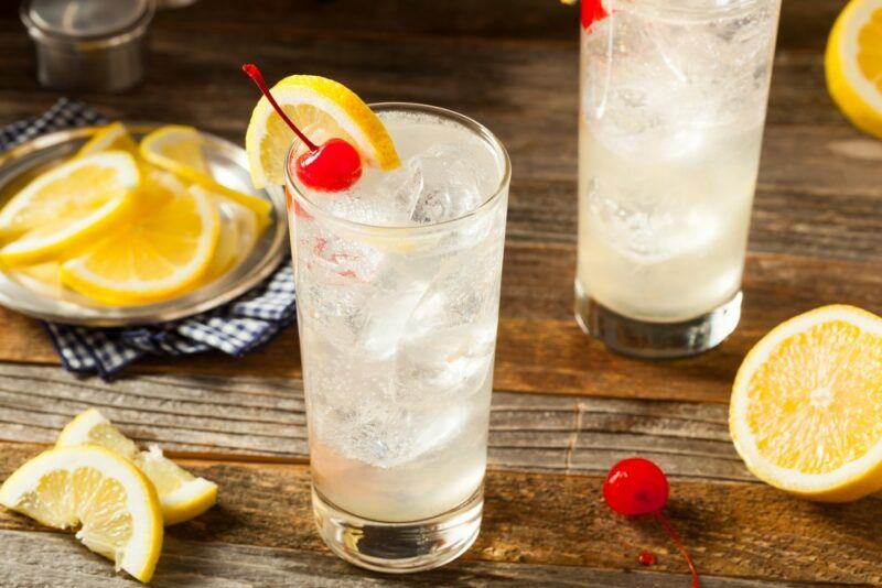 A wooden table with two tall glasses of a Tom Collins cocktail, next to some lemons