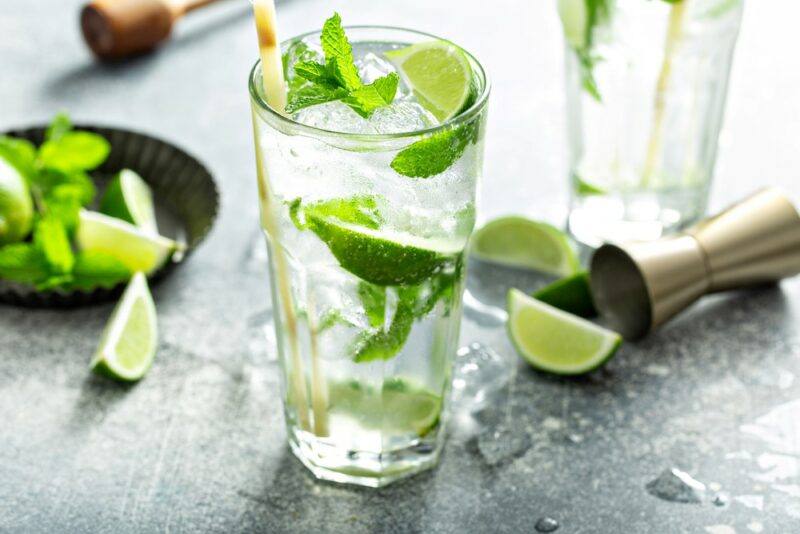 Two glasses of a White Claw mojito with lime and mint leaves, with more lime wedges on the table and in a bowl, plus a cocktail jigger