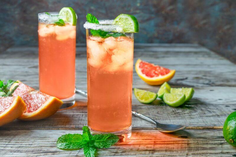 Two tall glasses containing a White Claw paloma cocktail with ice, mint leaves, lime, and a salt rim, with a grapefruit wedge, some lime wedges, a spoon, and peppermint leaves on the table