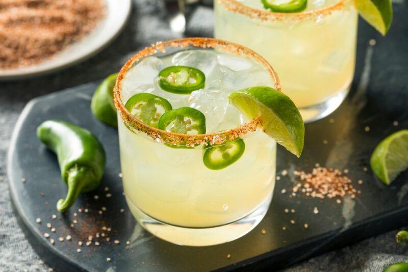 Two glasses containing a White Claw spicy margarita with jalapenos and a lime wedge as a garnish, with more jalapenos and some spicy salt on the table