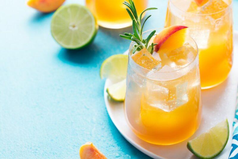 Two glasses of a basil peach moonshine cocktail on a white plate, with pieces of lime and peaches scattered around