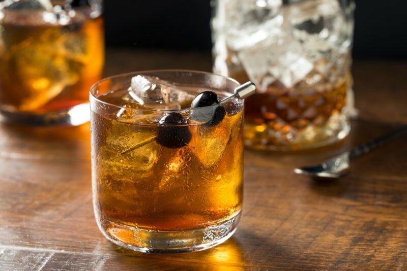 Two glasses containing a black cherry bourbon cocktail with plenty of ice and a cherry as a garnish, with a glass of whiskey and ice in the background