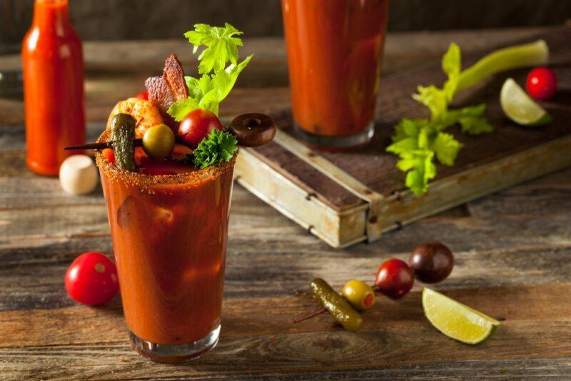 A single large glass of a Guinness bloody Mary on a table, with a large garnish, with plenty more garnishes on the table and a second cocktail in the background