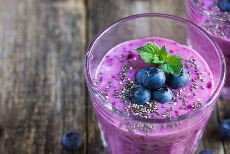 A glass with a brightly colored blueberry smoothie, with a second glass of the same smoothie just in the frame