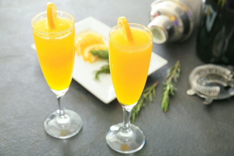 Two glasses of a bucks fizz cocktail in front of ingredients and a cocktail shaker