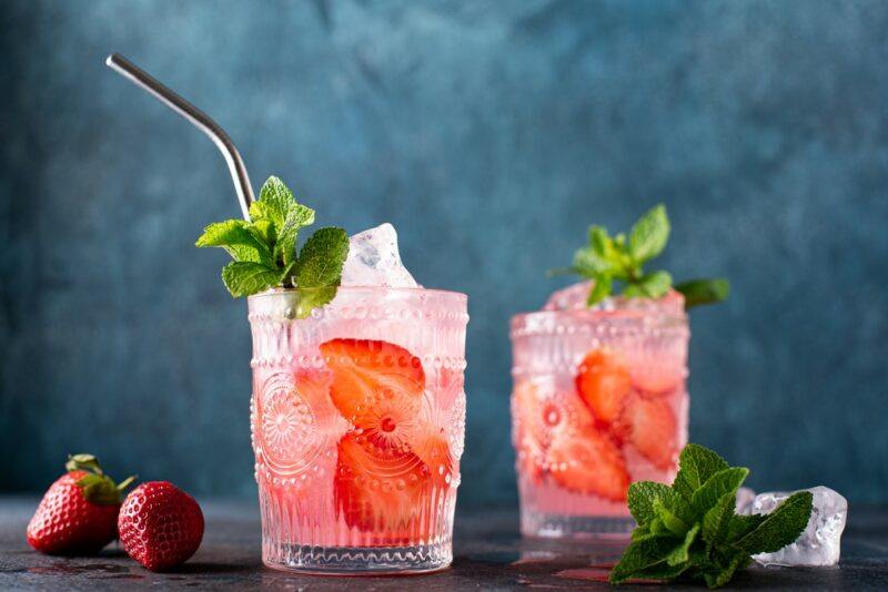 Two glasses of a cran strawberry blush cocktail, garnished with mint, one of which has a straw.