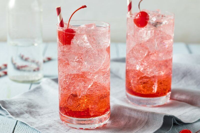 Two tall glasses containing a dirty Shirley Temple cocktail with ice and a cherry