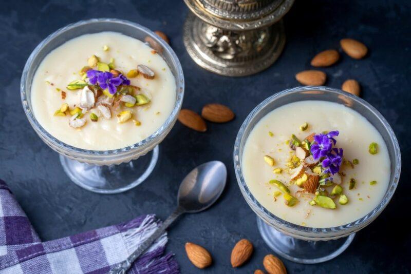 A dark table with two glasses of keskul dessert, with some almonds and a spoon on the table