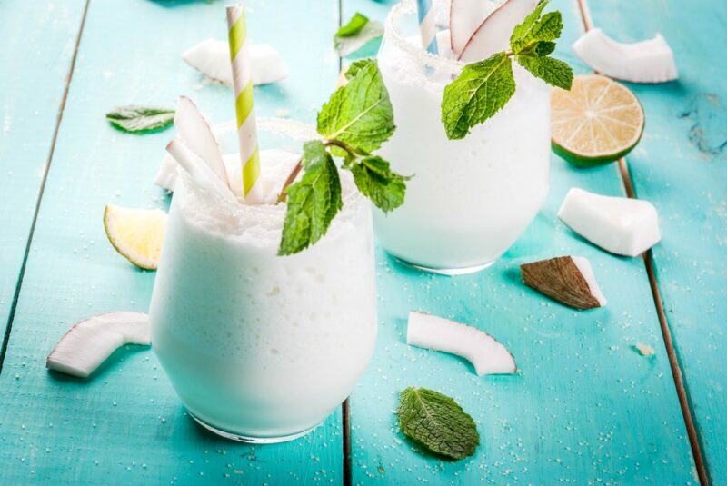 Two glasses containing a frozen toasted coconut cocktail on a light blue table with pieces of coconut and leaves scattered around