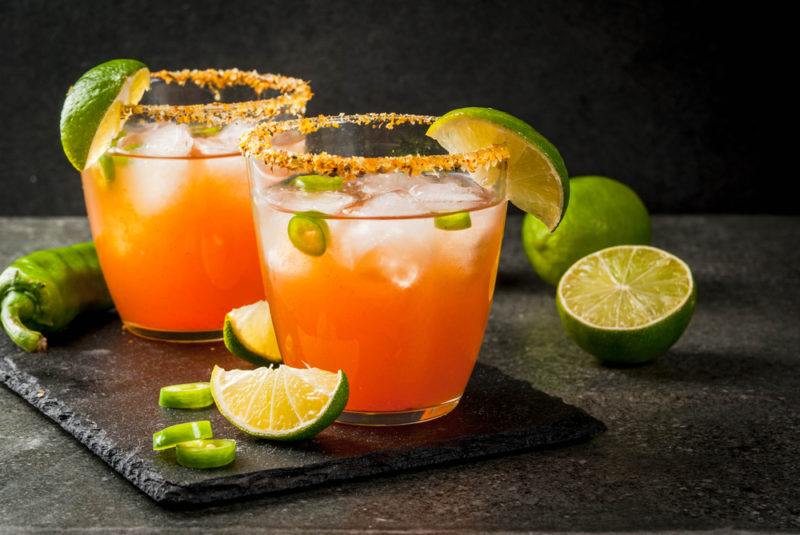 Two glasses of a ghost pepper sour cocktail with a spicy salt rum and limes on the table