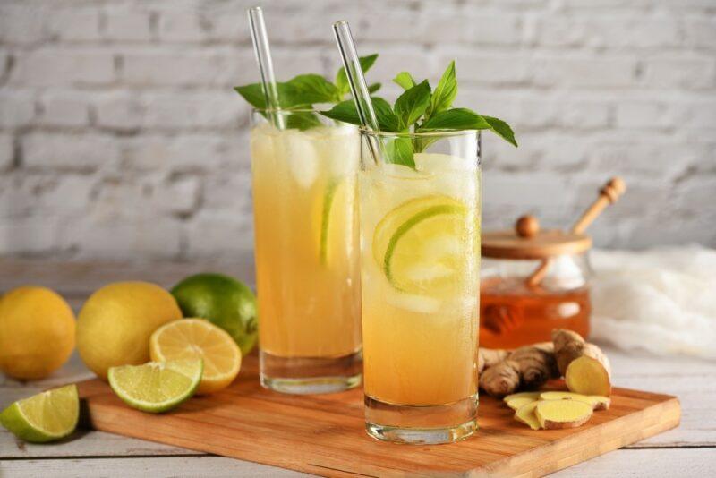 Two glasses containing a honey margarita with a straw, garnished with leaves, next to lemons, limes, and some honey