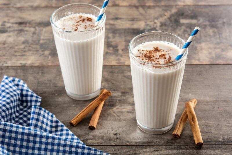 Two tall glasses containing a hot rum horchata cocktail with straws, next to cinnamon sticks and a blue and white cloth