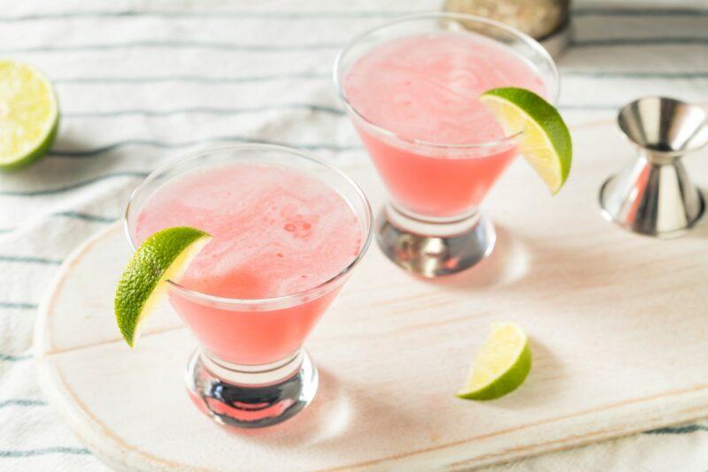 Two glasses containing a pink cosmopolitan, with lime wedges, next to a cocktail jigger