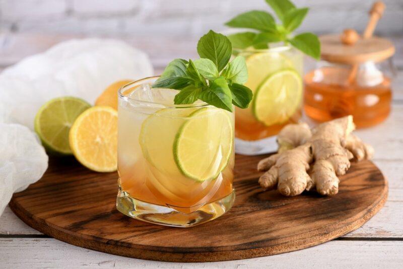 A wooden board with two glasses containing a mead cocktail, with sliced lime, next to ginger, limes, and some honey