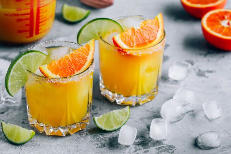 Two glasses containing a mimosa margarita, each garnished with an orange wedge and a slice of lime. There is ice and pieces of fruit scattered across the table