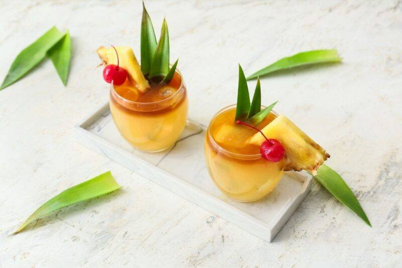 A white board with two glasses of a new moon mai tai cocktail, surrounded by pineapple leaves