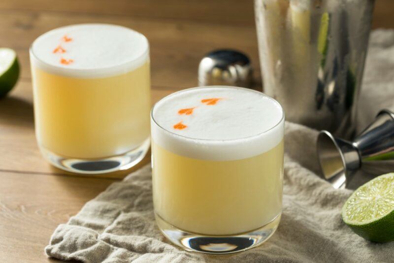 Two peanut butter whiskey sour cocktails. One is on a wooden table and the other is on a cloth, next to a cocktail shaker and a lime half