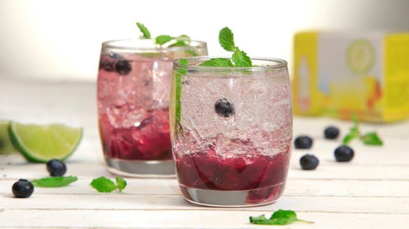 Two glasses of a peanut butter whiskey with blue Red Bull cocktail with plenty of crushed ice, next to some blueberries and leaves and a box of soda