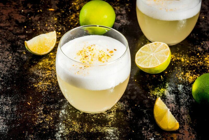 Two glasses containing a sake sour cocktail on a table with lemon and lime wedges