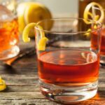 Two glasses containing neat sazerac cocktails with a lemon twist, next to a bottle of spirits and a lemon