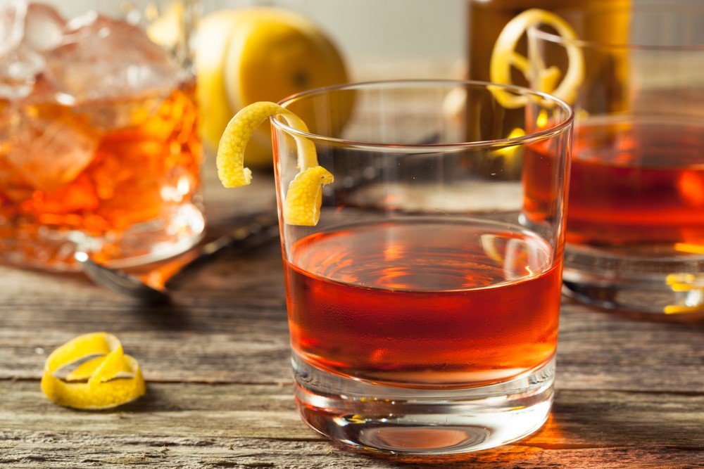 Two glasses containing neat sazerac cocktails with a lemon twist, next to a bottle of spirits and a lemon