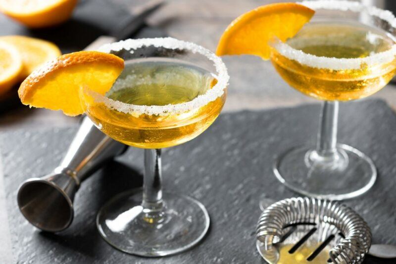 A piece of slate with two glasses of a sidecar cocktail, each rimmed with sugar and garnished with a orange wedge