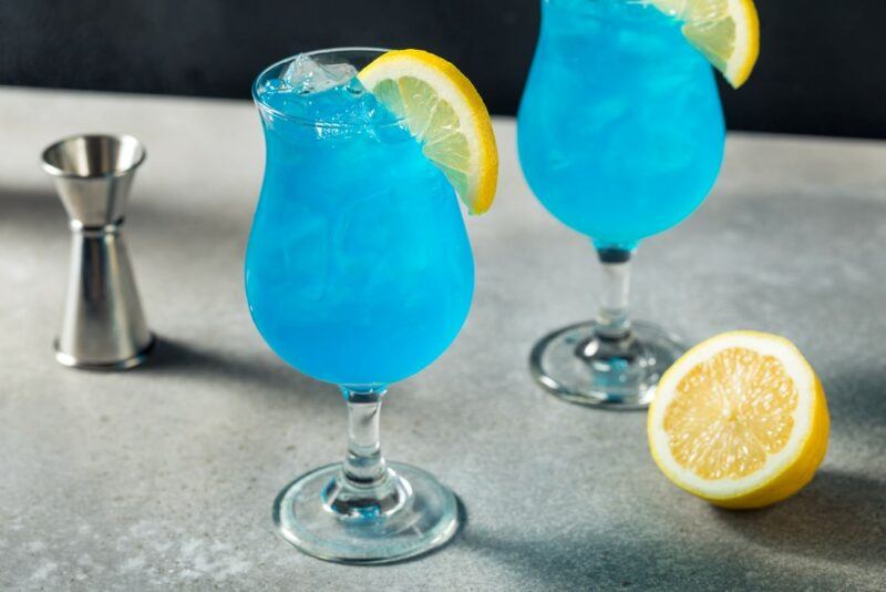 Two hurricane glasses containing a bright blue sour blue raspberry White Claw cocktail, each garnished with a lemon slice, with half a lemon and a cocktail jigger on the table