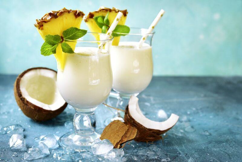 Two glasses containing a tropical paradise peanut butter whiskey cocktail, each garnished with a pineapple wedge and some greenery, with coconuts and ice on the table