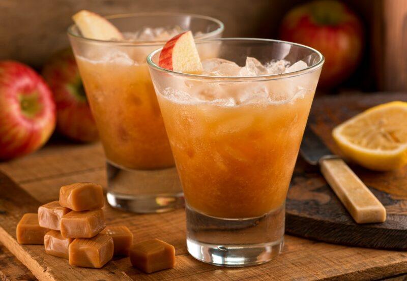 Two glasses containing an apple cider peanut butter whiskey cocktail with plenty of ice and apples as a garnish, there is caramel, apples, and oranges on the table