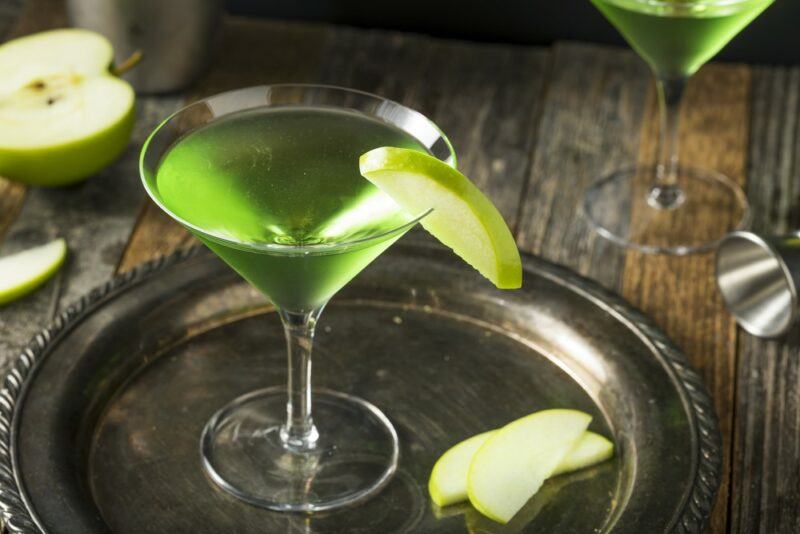 A silver tray with a glass of an appletini cocktail, with a second glass in the background and some apples