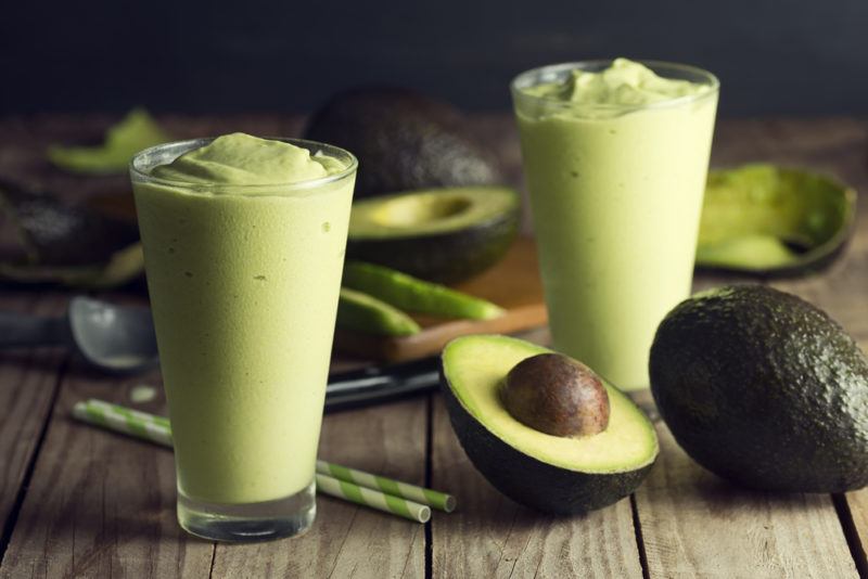 Two avocado smoothies in glasses, on a wooden table with avocados and straws