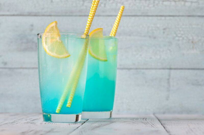 A white wooden table with two glasses of an electric lemonade cocktail with yellow straws