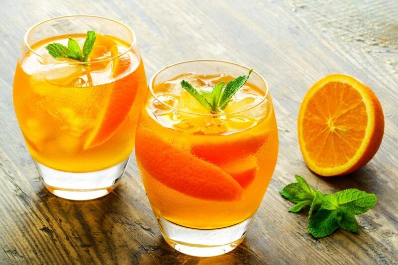 A wooden table with two glasses of an orange crescent cocktail, next to half an orange and some leaves