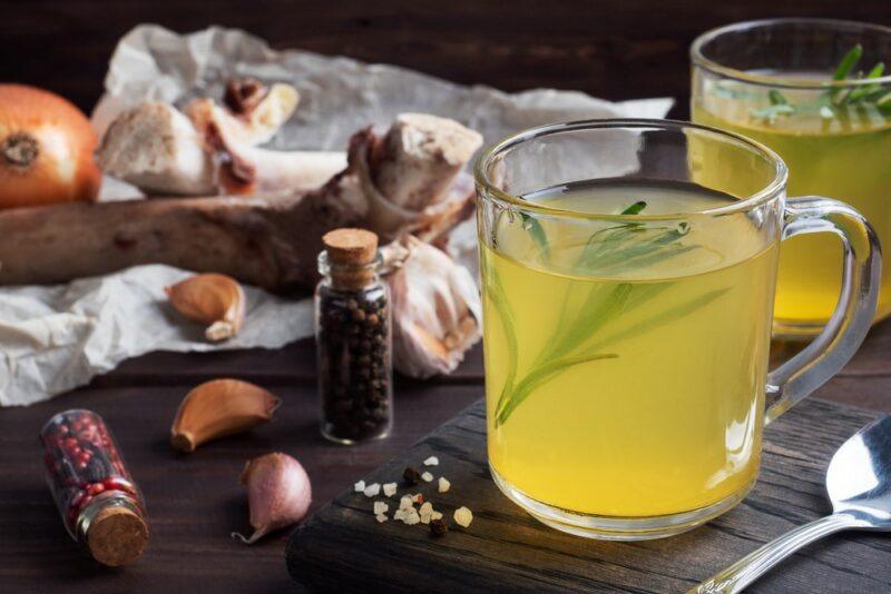 Two glasses of bone broth with rosemary, next to the ingredients to make bone broth