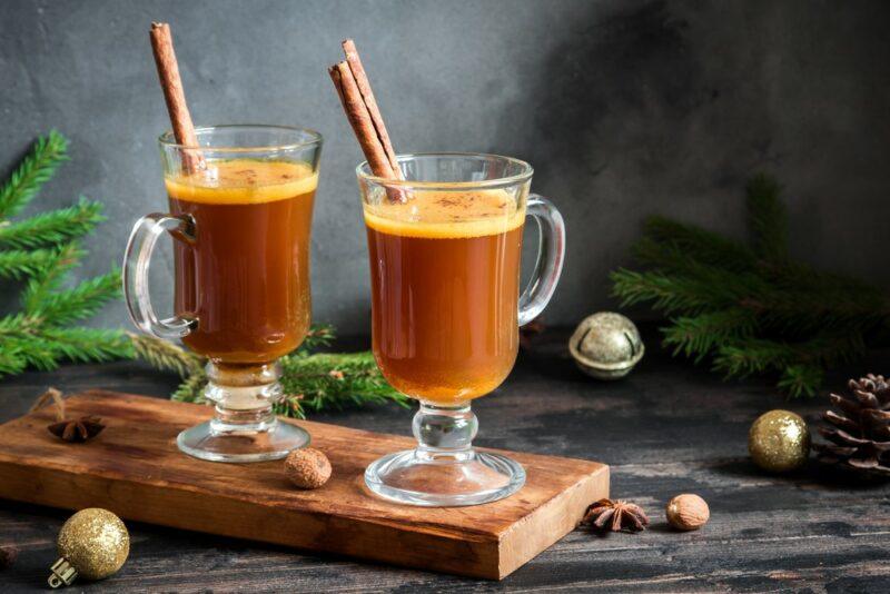 Two glass mugs of hot buttered rum with cinnamon sticks on a wooden board with Christmas decorations and pine tree fronds in the background