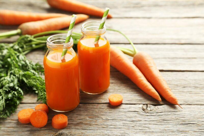 A wooden table with fresh carrots, a few carrot slices, and carrot juice