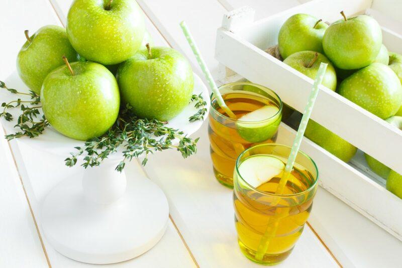 Two glasses of green apple juice on a white table near two stacks of green apples