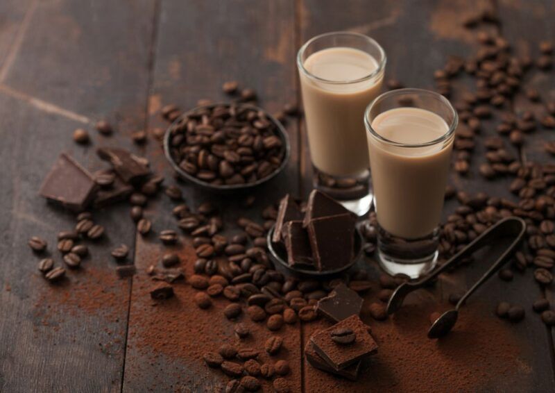 Two shot glasses containing a homemade Bailey's Irish Cream liqueur, surrounded by coffee beans and chocolate