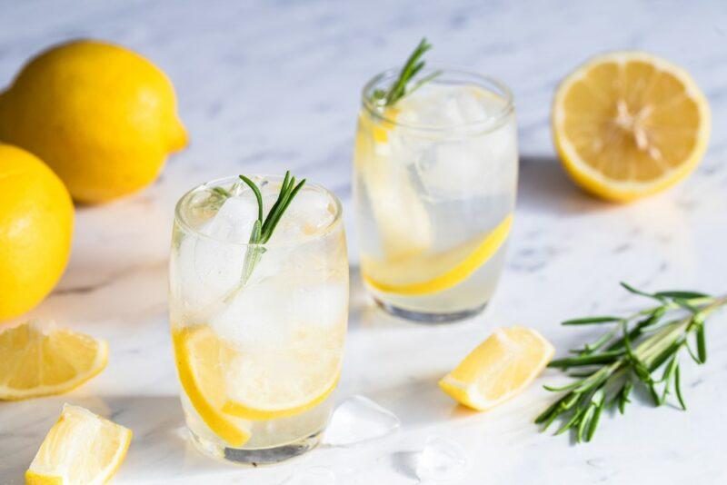 Two glasses of lemon and tonic water on a light colored table with lemons and rosemary nearby