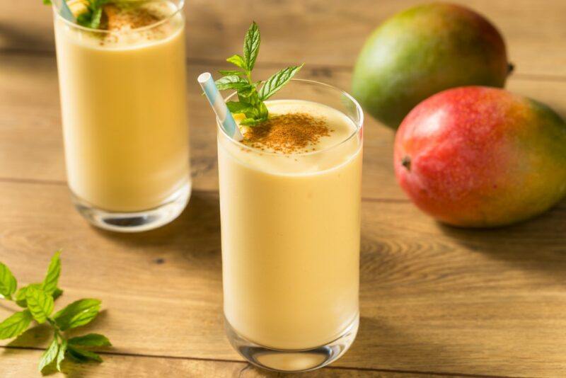 A wooden table with two glasses of mango lassi, next to a couple of mangos