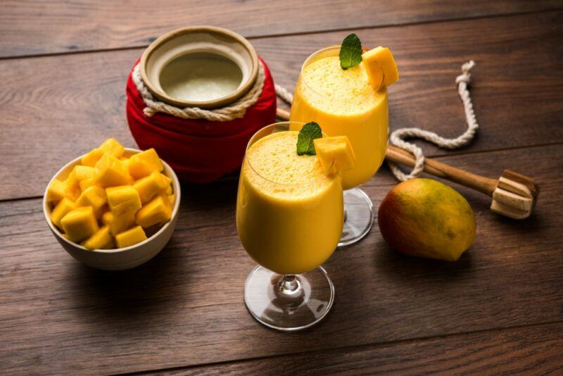 Two glasses of mango lassi on a wooden table, next to a pot and a bowl of mango pieces