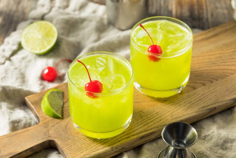 A wooden board with two melon patch cocktails with cherries. There is a lime segment on the board too, along with a cherry and a lime half in the background