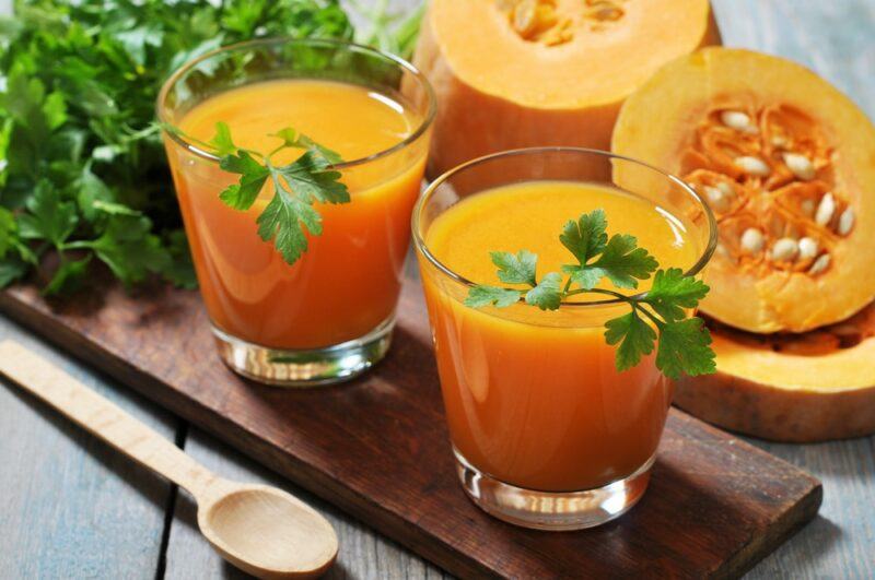 Two glasses of pumpkin juice next to pumpkin halves on a wooden board, next to a spoon