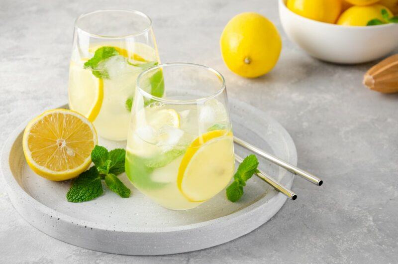 A white tray with two glasses of spiked lemonade, plus straws and half a lemon. There is a bowl of lemons in the background.