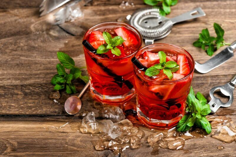 Two glasses containing a strawberry jam moonshine cocktail on a wooden table surrounded by ice